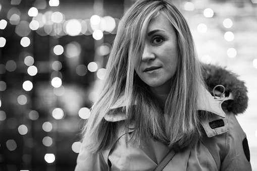 Winter portrait of young woman walking in snowy city decorated for Christmas and New Year holidays, wearing in a white coat with fur hood. Christmas lights on background. Black and white photo.