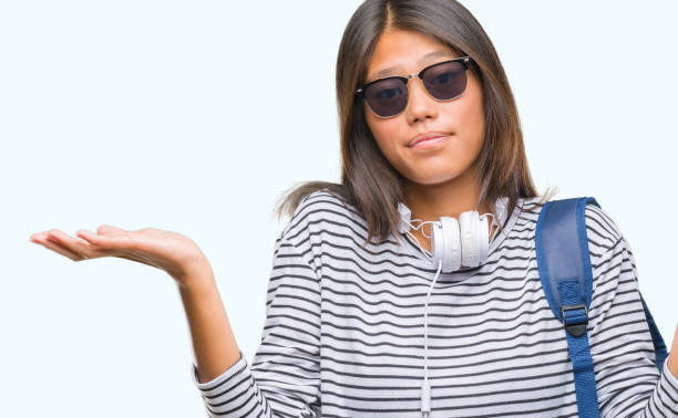 young asian student woman wearing headphones and backpack over isolated background clueless and confused expression with arms and hands raised. doubt concept. - head and shoulders audio imagens e fotografias de stock
