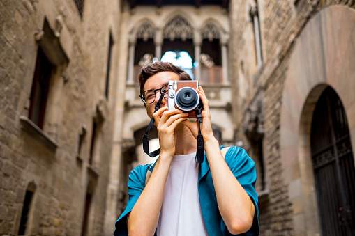 Young tourist photographing the Gothic quarter of Barcelona with an old camera. He is wearing casual clothes, eyeglasses, and a backpack.
