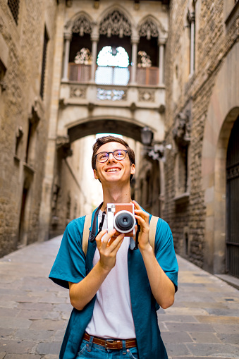 Young tourist photographing the Gothic quarter of Barcelona with an old camera. He is wearing casual clothes, eyeglasses, and a backpack.