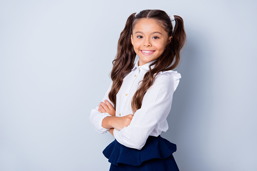 Welcome to first grade! Nice cute tender cheerful lovely small little girl with curly pony-tails in white formal blouse and blue skirt, folded arms, half-turned. Isolated over grey background