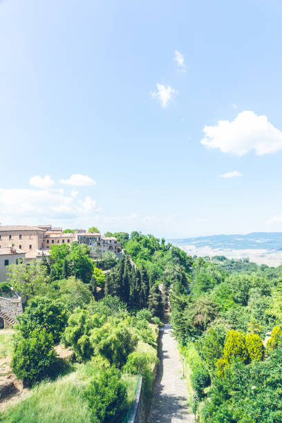 istock Vertical photo with empty road. The track goes among several green trees along ancient town Voltera. The town is in Tuscany - Italy. The sky is blue with few white clouds. 1056838230