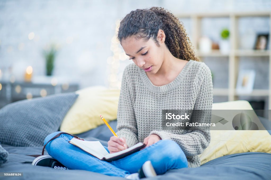Teen girl journaling on her bed A pretty teenaged girl writes into a journal with interest while sitting cross legged on her bed. Teenager Stock Photo