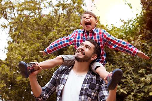 A father and his son are playing happily, while the boy is riding on his shoulders, smiling and playing both, on a beautiful afternoon in the park