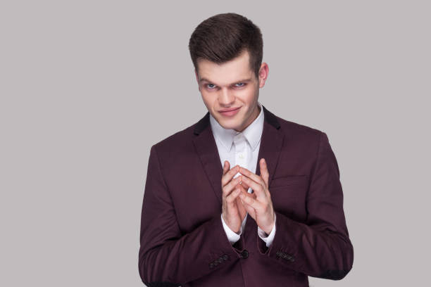 Portrait of funny cunning handsome young man in violet suit and white shirt, standing, looking at camera with cheating face. Portrait of funny cunning handsome young man in violet suit and white shirt, standing, looking at camera with cheating face. indoor studio shot, isolated on grey background. stealth stock pictures, royalty-free photos & images