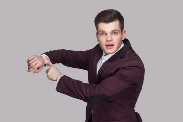 Photo of We have no more time. Portrait of handsome serious worry young man in violet suit and white shirt, standing, looking at camera and showing time gesture.