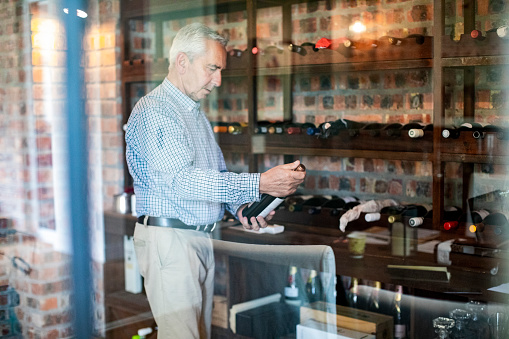 Senior man holding wine bottle while standing in cellar. Retired male is amidst wine collection at home. He is seen through glass.