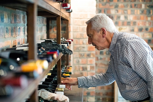 Side view of senior man choosing wine bottle from rack. Retired male is standing in cellar at home. He is in formalwear.