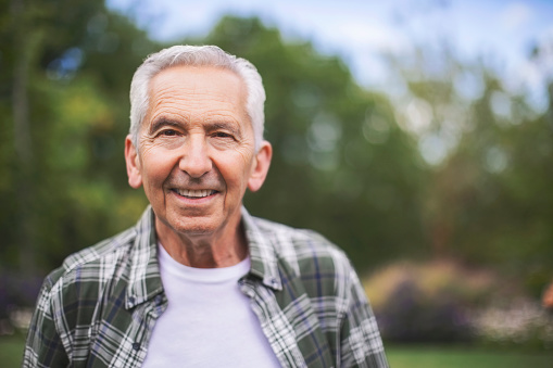 Portrait of smiling retired man standing in back yard. Close-up of happy senior male. He is in plaid shirt.