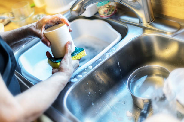 A woman is hand washing a coffee cup and other crockery dishes and glasses and cooking pots and pans using a yellow cleaning sponge. She is working in a two section stainless steel home kitchen sink.