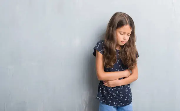 Photo of Young hispanic kid over grunge grey wall with hand on stomach because indigestion, painful illness feeling unwell. Ache concept.