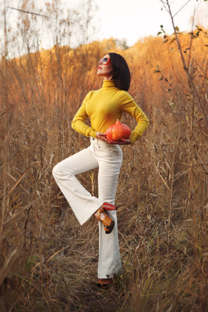 Fashionable beautiful woman in sunglasses standing in the field and holding a pumpkin for Halloween Fashionable beautiful woman in sunglasses standing in the field and holding a pumpkin for Halloween. She's wearing white bell bottom pants and yellow jacket flare pants stock pictures, royalty-free photos & images