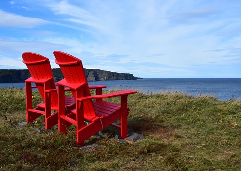two empty red Adirondack chairs on the edge of a cliff overlooking the ocean and coastline, Silver Mine Head Path Torbay Newfoundland Canada