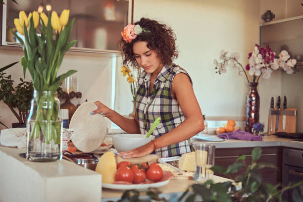 urocza kręcona latynoska dziewczyna gotuje w swojej kuchni. - stereotypical housewife women domestic kitchen brown hair zdjęcia i obrazy z banku zdjęć