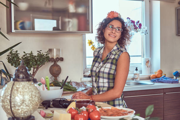 urocza kręcona latynoska dziewczyna gotuje w swojej kuchni. - stereotypical housewife women domestic kitchen brown hair zdjęcia i obrazy z banku zdjęć