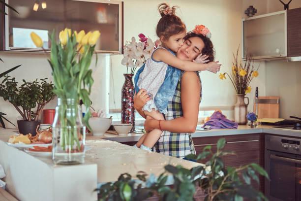 słodka dziewczynka całuje swoją mamę, siedząc w ramionach w kuchni. - stereotypical housewife little girls family domestic kitchen zdjęcia i obrazy z banku zdjęć