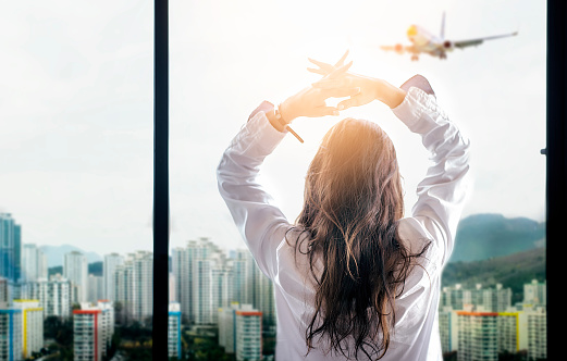 Rear view of woman with long hair standing at window side and looking at city view while the airplane fly over