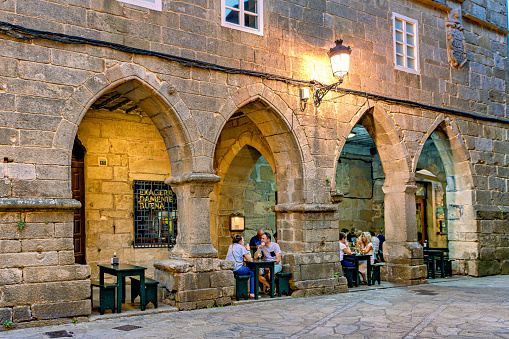 Tourists and locals enjoy an evening drink in outdoors bar in historic stone building in old town Noia, Galicia, Spain.