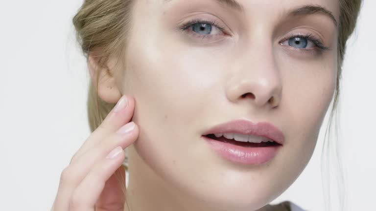 Close-up of smiling woman applying face cream. Portrait of young female is taking skin care against white background. She is having blue eyes.