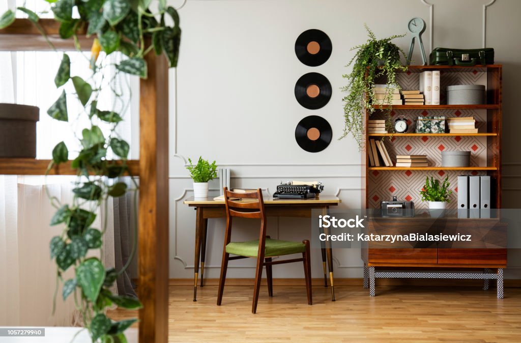 Black retro typewriter on a unique wooden desk, a mid-century modern chair and a renovated bookcase in a hipster home office interior. Real photo. Retro Style Stock Photo