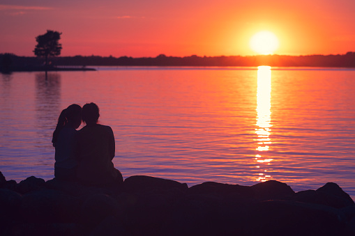 A mother and daughter sits closely together by a lake at sunset watching the sun go down.