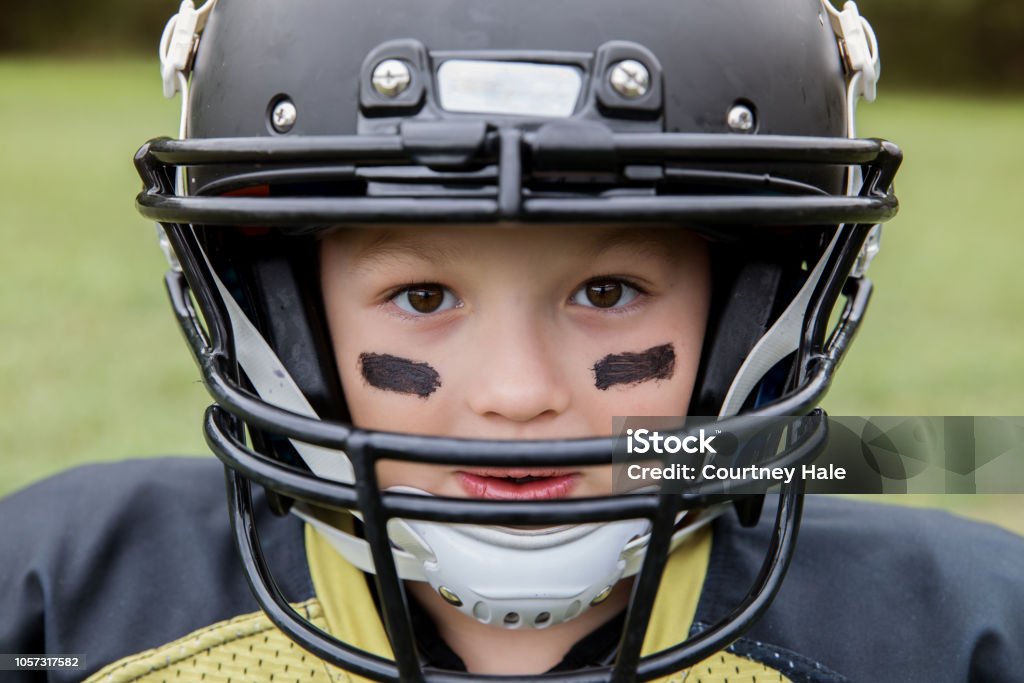 Garçon est petit joueur de football américain regarder dans la caméra à travers casque - Photo de Football américain libre de droits