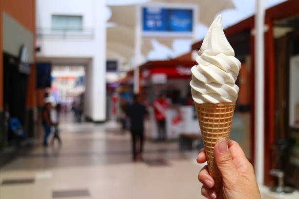 istock Woman's hand holding vanilla milk soft serve ice cream cone in the sunlight, with blurred shopping mall in background 1057323552