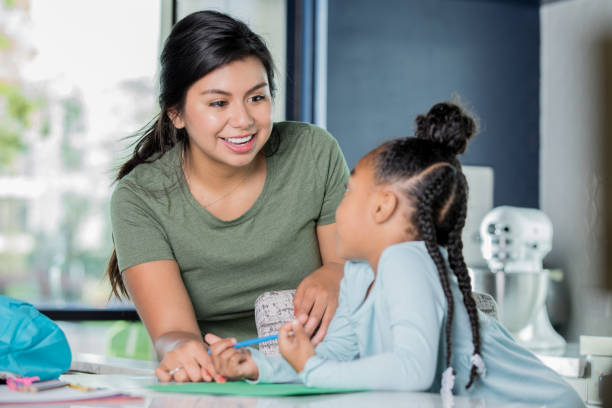 Happy young Hispanic woman is babysitting, tutoring elementary age girl. Young adult Hispanic woman is smiling while babysitting and tutoring African American little girl. Nanny is assisting child with her homework in modern kitchen. nanny stock pictures, royalty-free photos & images