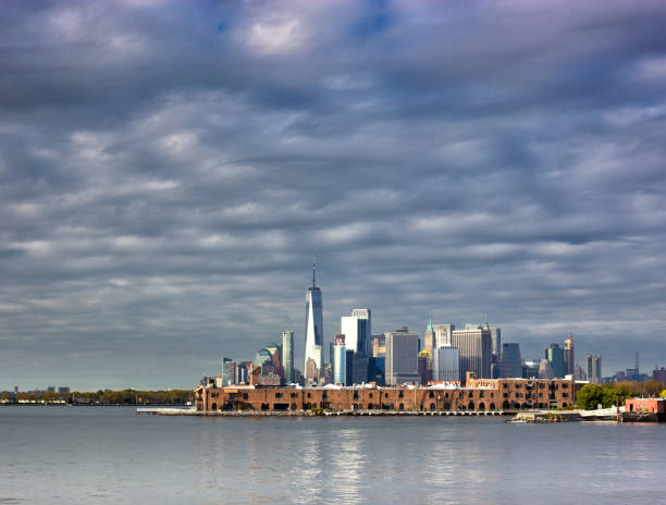 New York, NY: A sunlit Lower Manhattan/Wall Street skyline as seen across the New York Harbor from Red Hook, against a dramatic sky. Copy space available at the top or bottom of the frame (sky or river).