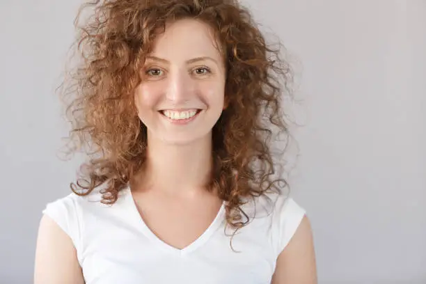 Headshot Young cheerful happy curlyhair ginger girl smiling broadly and looking at camera over gray background. European shaggy hirstyle female have good mood and pleasant face. People concept