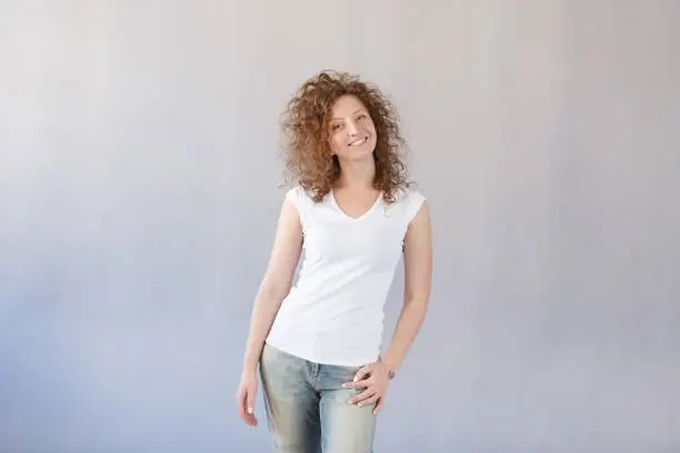 Young cheerful happy curly hair ginger girl smiling broadly and looking at camera over gray background. European redhead hirstyle female have good mood and pleasant face. People concept