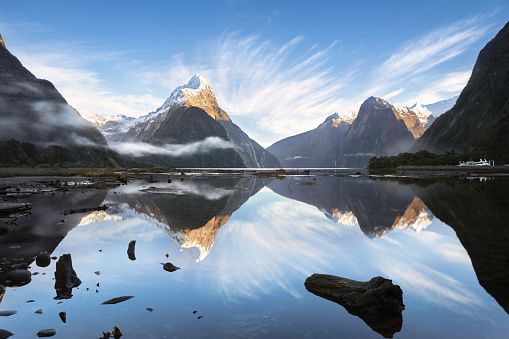 Early sun rays strike the mountain peaks at Milford Sound, New Zealand.  Streaky white cloud stretches out from behind the mountains. The still waters of the fjord reflect the scene.\nMilford Sound is situated in Fiordland National Park, on the south west of New Zealand's South Island. It is known for its shear cliffs rising straight out of the sea. Mitre Peak (centre left) is a famous landmark and rises 5,560 feet (1,690m) from sea level.