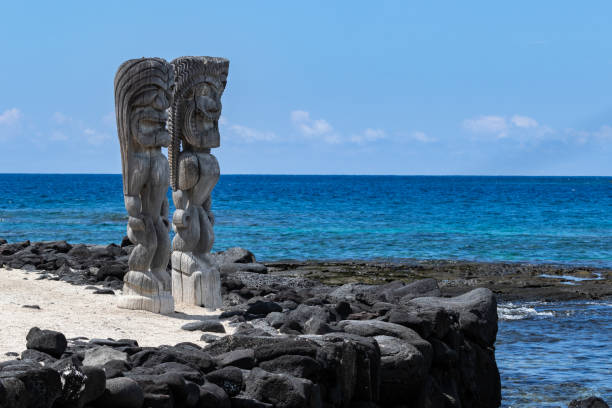Pair of wooden status (Tikis), place of refuge (Honaunau), Hawaii. Standing on white sand, next to barrier wall of black lava rock. Blue Pacific ocean & sky in background. Two Wooden Tikis in the place of refuge (Honaunau) sanctuary, on the Big Island of Hawaii. Standing in white sand with a black lava barrier wall; Pacific ocean and blue sky are in the background. big island hawaii islands photos stock pictures, royalty-free photos & images