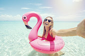 Young woman takes selfie portrait on idyllic beach with inflatable pink flamingo in pristine clear water in the Islands of Thailand. People travel luxury fun and cool attitude concept
