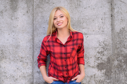 Woman smiling at camera. Woman with blonde hair, dressed in red checkered shirt and jeans standing against grey wall. Portrait, close up photo