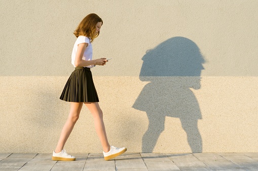 Young girl in the profile, walks along the gray wall, in the hands of mobile phone. Outdoor, copy space, golden hour