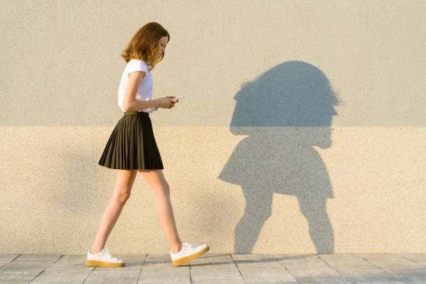 молодая девушка в профиле, ходит вдоль серой стены, в руках мобильного телефона. открытый, копия пространства - walking girl стоковые фото и изображения
