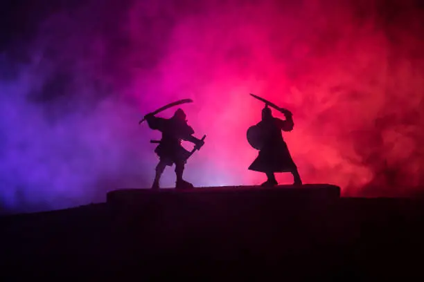 Silhouette of two medieval warriors in duel. Picture with two fighter with sword with dark toned foggy background. Selective focus.