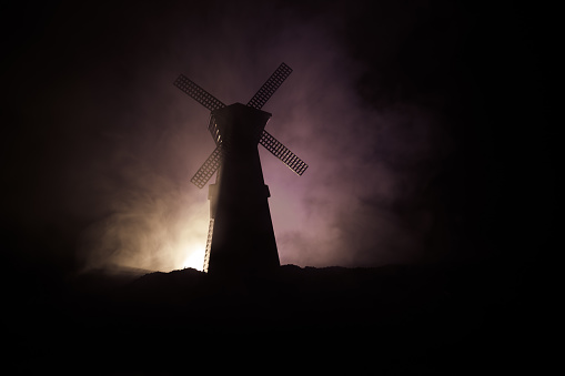 Windmill silhouette standing on hill against the night sky. Night decor with old windmill on hill with horror toned foggy background with light. Horror concept