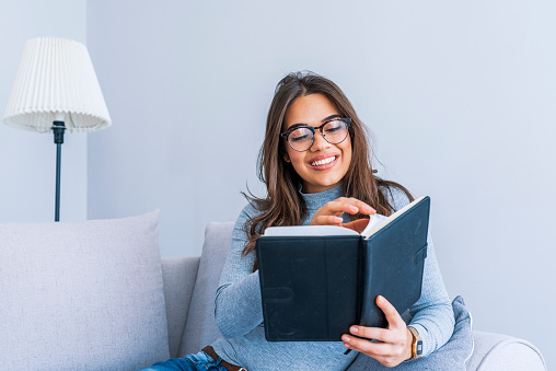 Excellent book. Beautiful young woman reading a book at the sunny morning. Happy young woman reading storybook on couch at home. Brunette on couch with book