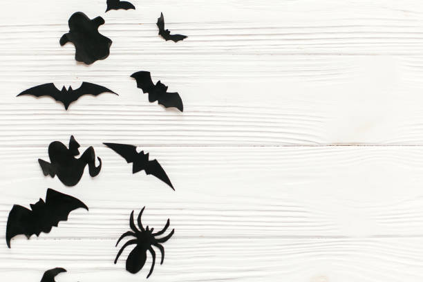 istock Halloween flat lay.  Black paper bats, spiders, ghosts top view on white rustic wooden background. Trick or treat. Space for text. Season's greeting card mockup. 1057539010
