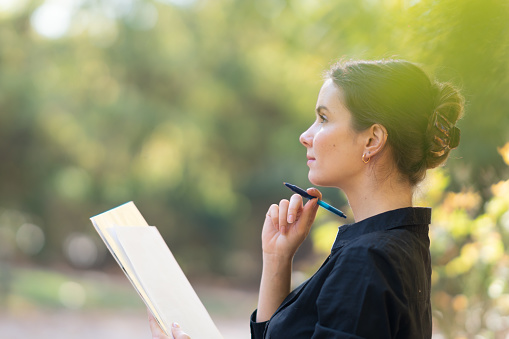 Young woman sitting on a bench out in the park holding a pen and papers, concentrating on text and notes she's working on.