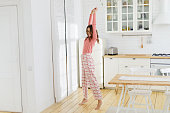 Happy young smiling girl dancing in bright white sunny kitchen at home, wearing pink checkered pajama pants