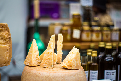 Horizontal color image depicting wedges of parmesan (Parmigiano Reggiano) on display and for sale at a street food market in London, UK. In the background glass bottles of fresh extra virgin olive oil are defocused, allowing room for copy space.