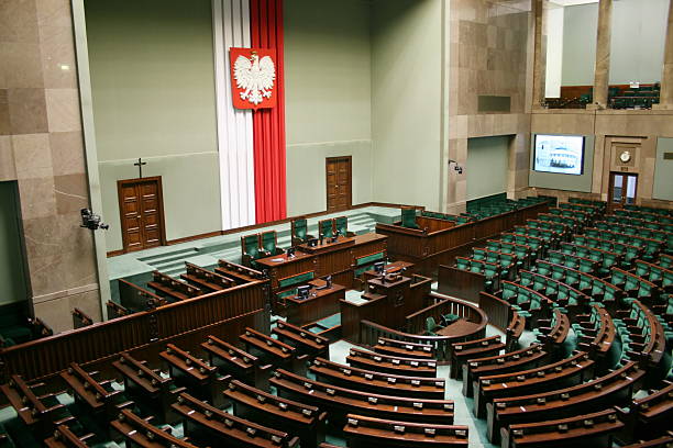 Polish parliamentary chamber with rows of green seats, a podium, and a large white eagle emblem on a red and white banner.