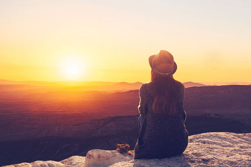 A view from the back of a girl in a hat sits on a hill and admires the beautiful view.
