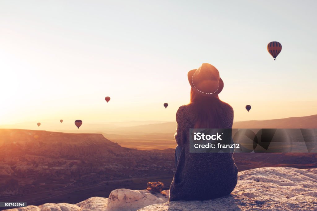 Vista desde la parte de atrás de una niña en un sombrero se asienta en una colina y mira los globos de aire. - Foto de stock de Viajes libre de derechos