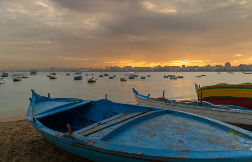 Turquoise blue fishing boat on the beach at sunrise with Alexandria skyline in far distance and colorful sky at sunrise, Egypt