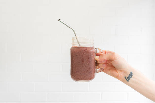 Woman's hand holding berry smoothie in glass mug with metal straw Woman's hand with love tattoo on wrist holding berry smoothie in glass mug with metal straw against white background  (selective focus) wrist tattoo stock pictures, royalty-free photos & images