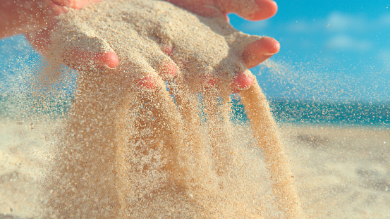 CLOSE UP: Unknown young woman scattering hot white sand between her gentle fingertips. Playful female tourist scooped up a big handful of sand and lets it get blown away in the cool ocean breeze.